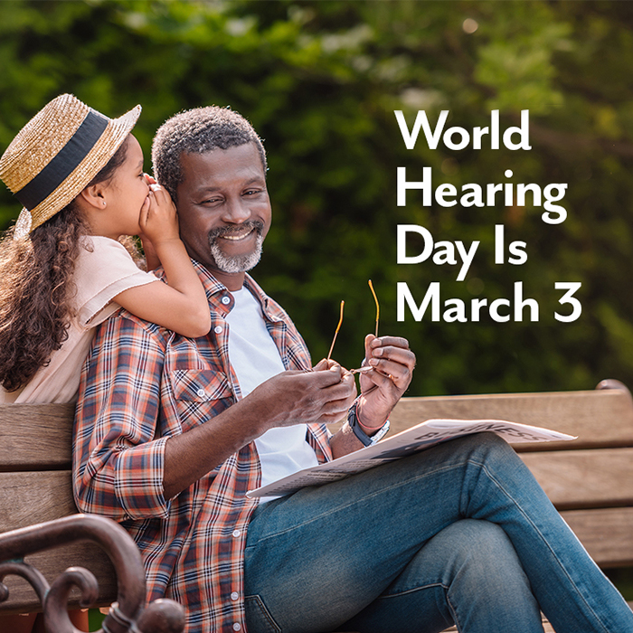 Young girl whispering to her grandfather who is sitting on a park bench. Text: World Hearing Day Is March 3