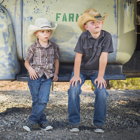 Two young boys wearing cowboy hats leaning against an antique truck in a rustic country setting.