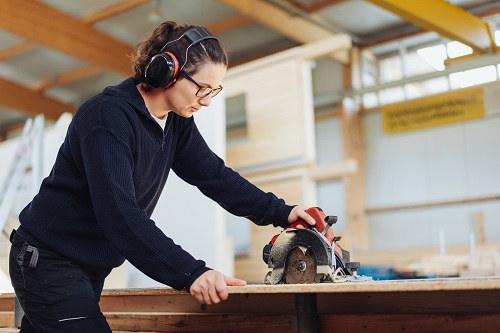 A woman uses a circular saw while wearing protective earmuffs.