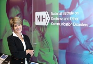 An image of Dr. Tucci standing in a hallway next to a sign that reads, "NIH National Institute on Deafness and Other Communication Disorders"
