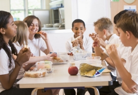 Kids sitting around a table eating lunch and talking to one another.